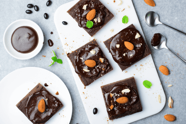Five healthy, sugar-free brownies topped with chocolate sauce and almonds on a white chopping board.